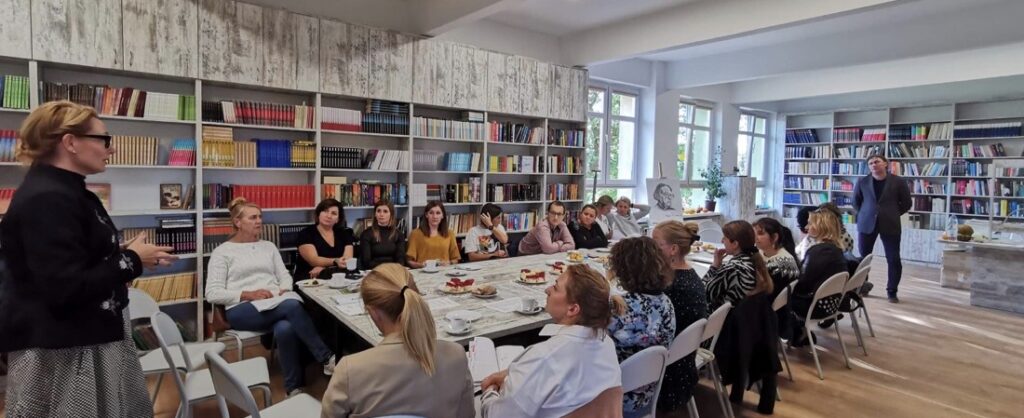 Sala biblioteczna, na środku stoły ustawione w kształcie prostokąta. Wokół siedzą nauczyciele. Za nimi stoi na tle regałów z książkami mężczyzna. Po prawej stronie regały z książkami. Na stole stoją talerzyki z ciastem i białe filiżanki. Przed stoi kobieta i coś mówi.

