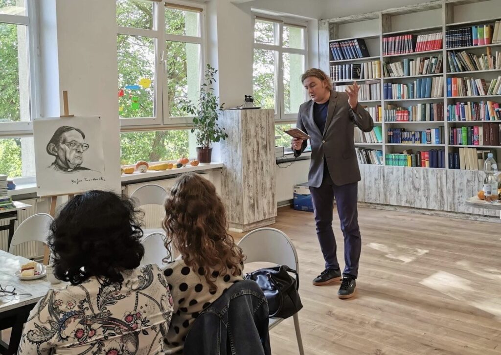 Sala biblioteczna. Na środku stoi mężczyzna i trzyma w ręku książkę. Za nim regały z książkami mężczyzna. Po prawej stronie regały z książkami. 