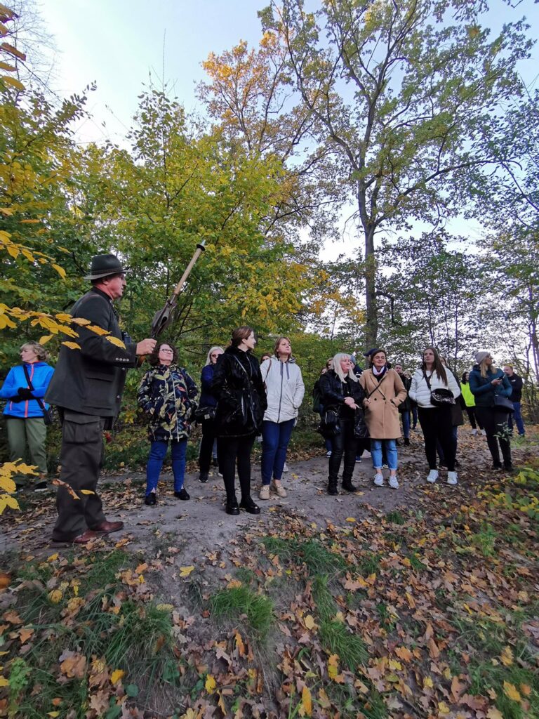 Na pierwszym planie stoi mężczyzna. Za nim duża grupa osób. Są w otoczeniu zielonych i żółtych drzew. Mężczyzna wskazuje na coś laską