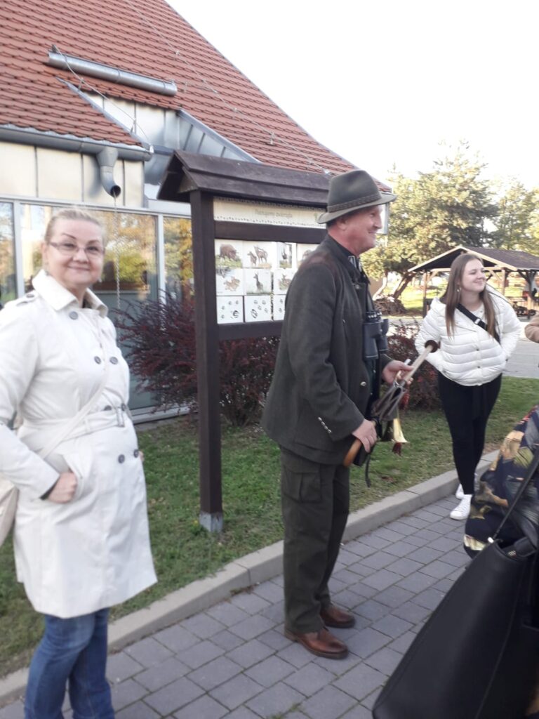 Na tle budynku stoi mężczyzna w Przed obiektem Regionalnego Ośrodka Edukacji Ekologicznej w Dziewinie stroju leśnika, po jego prawej i lewej stronie stoją dwie kobiety ubrane na jasno. Za nimi tablica poglądowa.