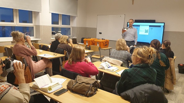 Na zdjęciu widoczna grupa nauczycieli, siedzą w ławkach. Mają założone okulary VR i patrzą przez nie. Z przodu stoi mężczyzna, który tłumaczy grupie użycie okularów. 