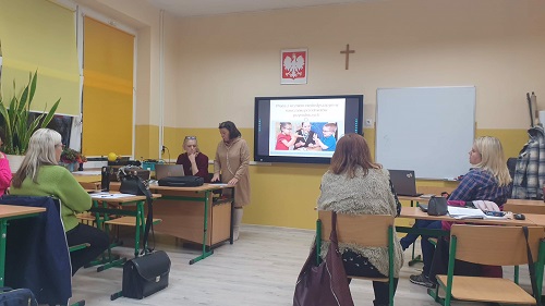 Sala lekcyjna, przy stolikach siedzą nauczyciele. Na ścianie ekran z prezentacją. Przed siedzącymi osobami znajdują się dwie kobiety prowadzące szkolenie. Jedna kobieta ubrana jest w bordową bluzką, druga w brązową sukienkę. Po lewej stronie okna przysłonięte żółtymi roletami. Po prawej stronie ekranu wisi biała tablica.