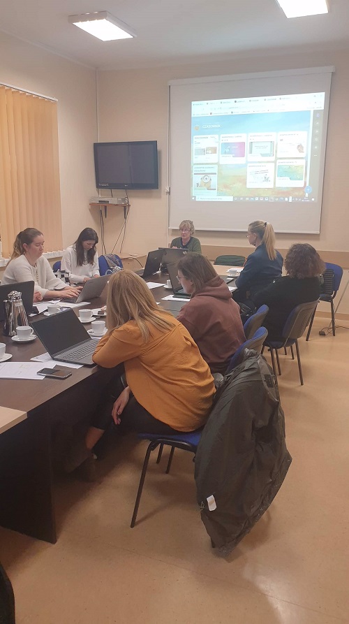 Sala szkoleniowa, przy stolikach siedzą nauczyciele. Na ścianie ekran z prezentacją. Przy stole, z przodu siedzi prowadząca, ma przed sobą otwartego laptopa.