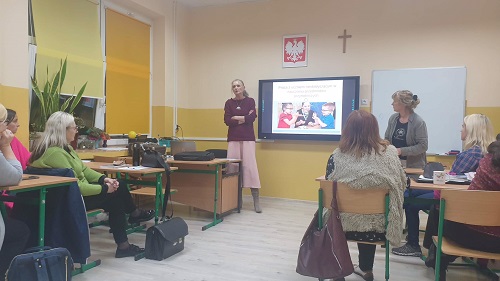 Sala lekcyjna, przy stolikach siedzą nauczyciele. Na ścianie ekran z prezentacją.. Po lewej stronie stronie ekran z prezentowanym materiałem a po jego prawej stronie wisi biała tablica. Przed ekranem stoi kobieta prowadząca ubrana w bordowy sweter i zieloną spódnicę, Przed ekranem druga prowadząca cos tłumaczy.