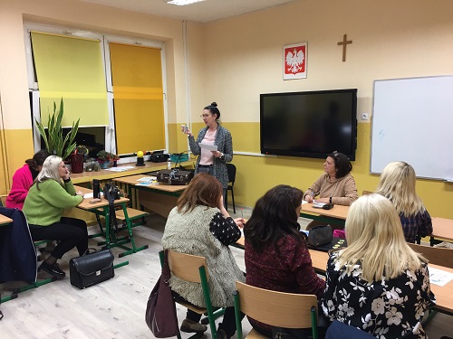 Sala lekcyjna, przy stolikach siedzą nauczyciele. Na ścianie ekran z prezentacją. Przed nimi stoi prowadząca i cos wskazuje ręką i tłumaczy. Ubrana jest w różową bluzkę i ciemny sweter. Po lewej stronie okno. Po prawej stronie ekranu wisi biała tablica.