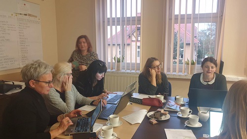 Sala szkoleniowa, przy stolikach siedzą nauczyciele i mają przed  sobą otwarte laptopy.Za nimi duże okna.