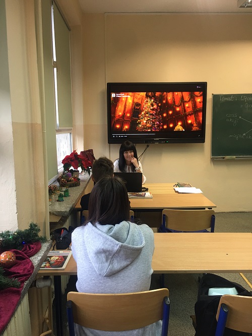 Sala lekcyjna, z przodu ekran z kolorową prezentacją, po jego prawej stronie zielona tablica do pisania. Przed monitorem siedzi przy biurku kobieta w białej bluzce. W ławce, przed nią siedzą uczniowie.