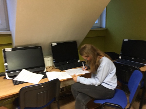 Sala multimedialna. Przy stoliku z komputerem siedzi i pisze kobieta w białym sweterku.