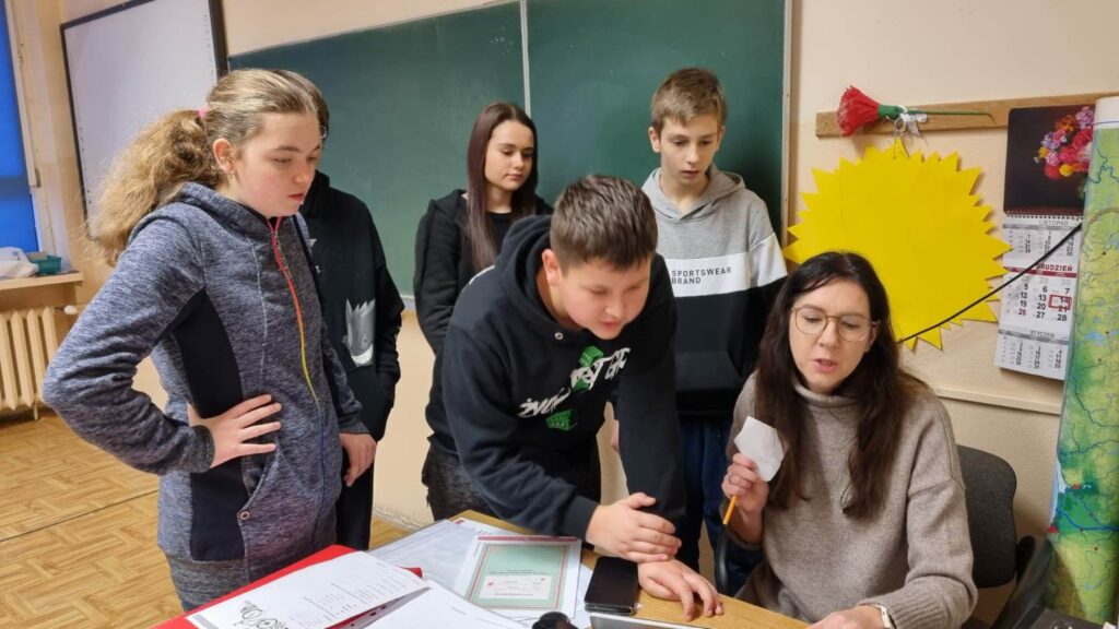 Nauczycielka siedzi przy biurku, kilkoro uczniów stoi koło niej. 