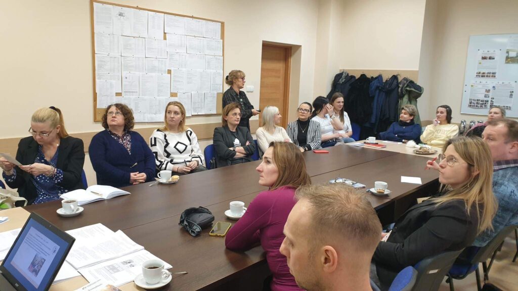 Sala szkoleniowa, przy stolikach siedzą nauczyciele. Za siedzącymi osobami, na tle ściany stoi kobieta prowadząca szkolenie. 