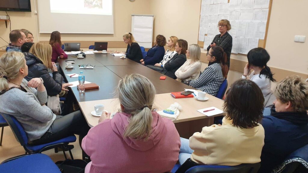 Sala szkoleniowa, przy stolikach siedzą nauczyciele. Na ścianie biała tablica informacyjna. Na ścianie z przodu ekran z prezentacją. Po prawej stronie stoi kobieta prowadząca szkolenie.