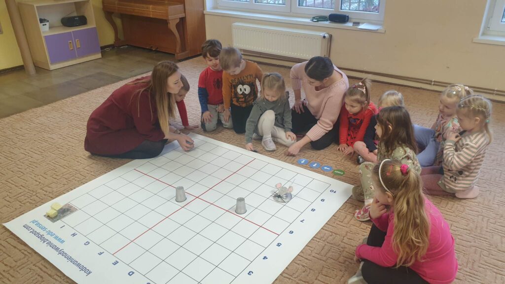 Dzieci i Panie siedzą na dywanie. Przed nimi leży mata do kodowania, na macie dwa szare kubeczki i myszka zrobiona z kartonu. 
