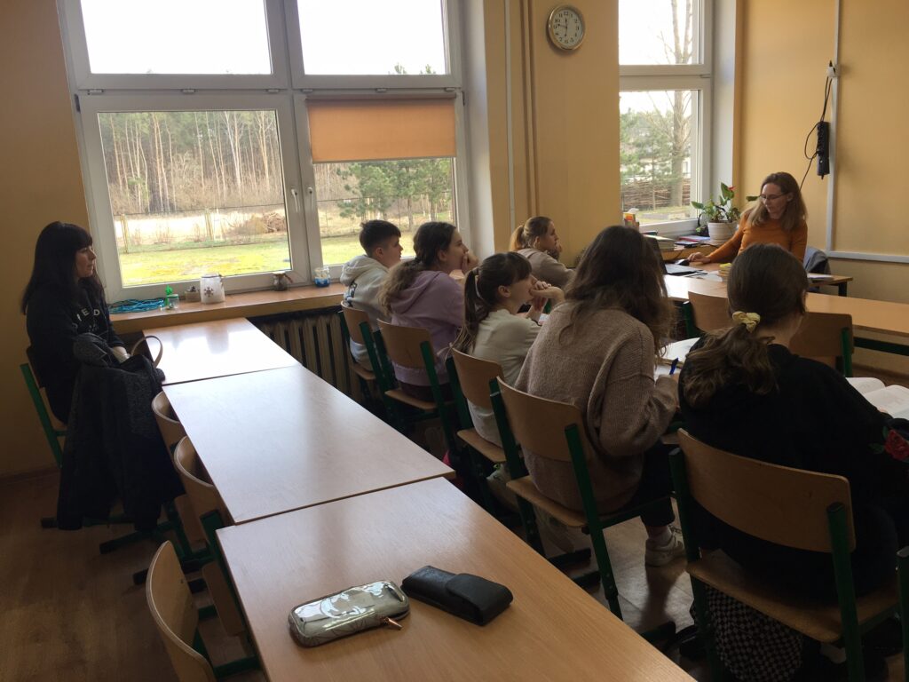 Sala lekcyjna. Na środku uczniowie w ławkach, siedzą twarzami do tablicy. Po lewej stronie siedzi przy biurku nauczyciel prowadzący zajęcia. Z tyłu siedzi dorosły obserwator.