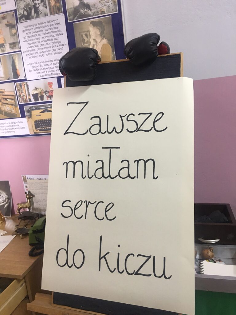 Na środku sztaluga z czarnym napisem na białym tle. Na górze sztaluga rękawice bokserskie.