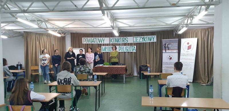 Na zdjęciu widać  7 kobiet oraz uczestników konkursu powiatowego. Za nimi napis Powiatowy Konkurs Młody Poliglota, baner Powiatowego Centrum Edukacji i Pomocy Psychologiczno- Pedagogicznej w Wołowie  oraz baner Miejsce Odkrywania Talentów -  Powiatowe Centrum Edukacji i Pomocy Psychologiczno- Pedagogicznej w Wołowie . Na każdej ławce stoi butelka wody mineralnej.  
