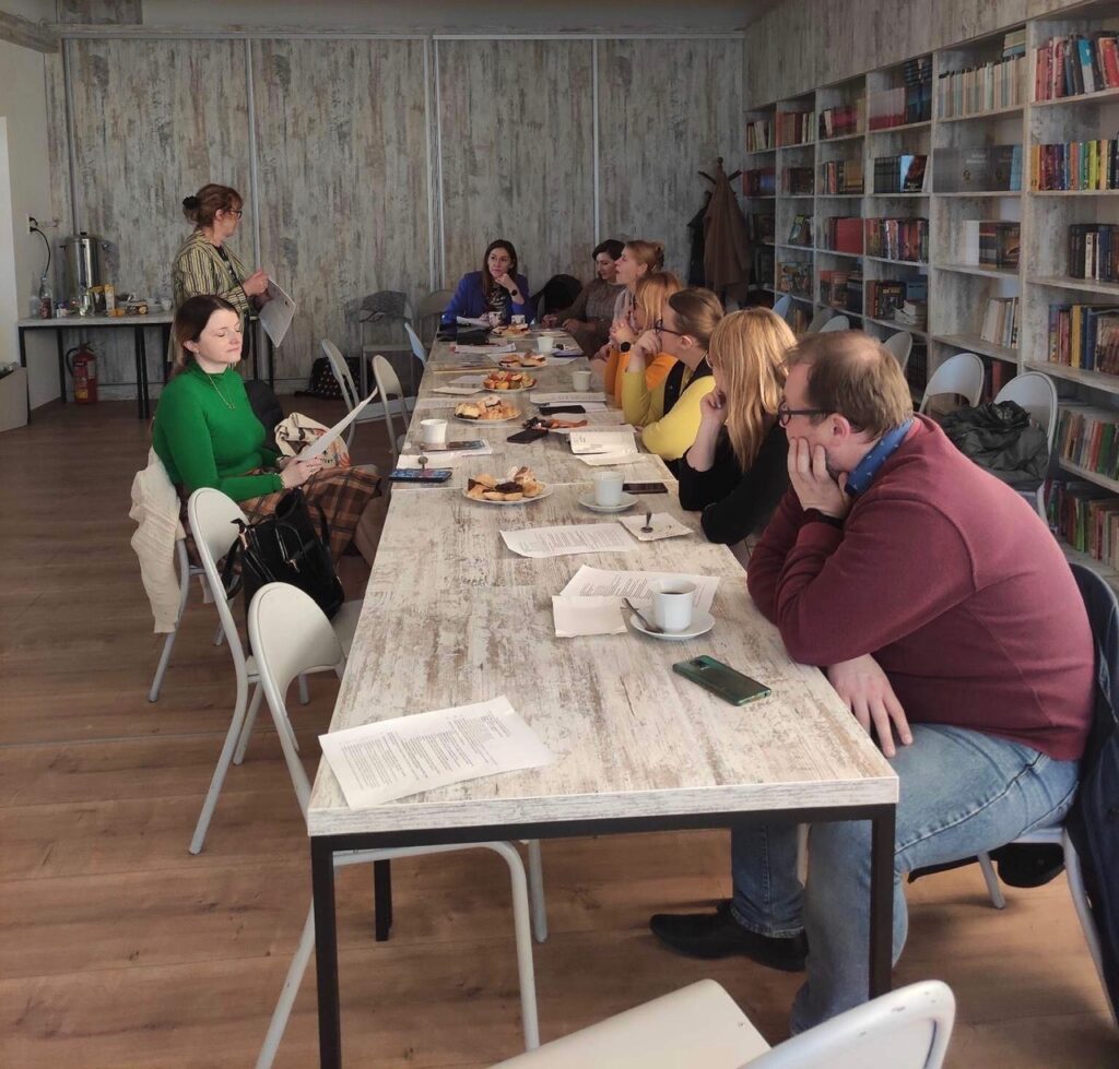 Sala biblioteczna, na środku stoły ustawione w kształcie prostokąta. Wokół siedzą nauczyciele. Z boku stoi kobieta i coś mówi. Po lewej stronie regały z książkami a z przodu ekran z wyświetlonym materiałem. Na stole stoją talerzyki z ciastem i białe filiżanki