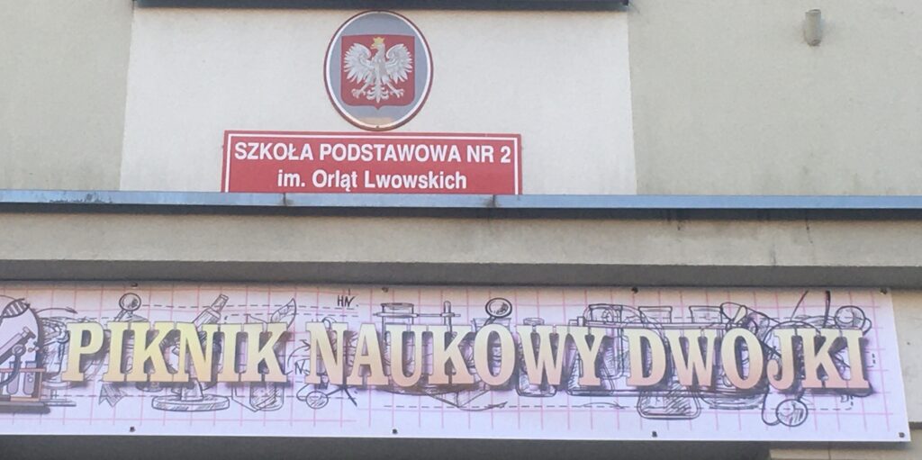 Budynek, na którym widnieje na czerwonym tle biały napis- nazwa szkoły. Poniżej kolorowy baner z napisem „Piknik Naukowy Dwojki”