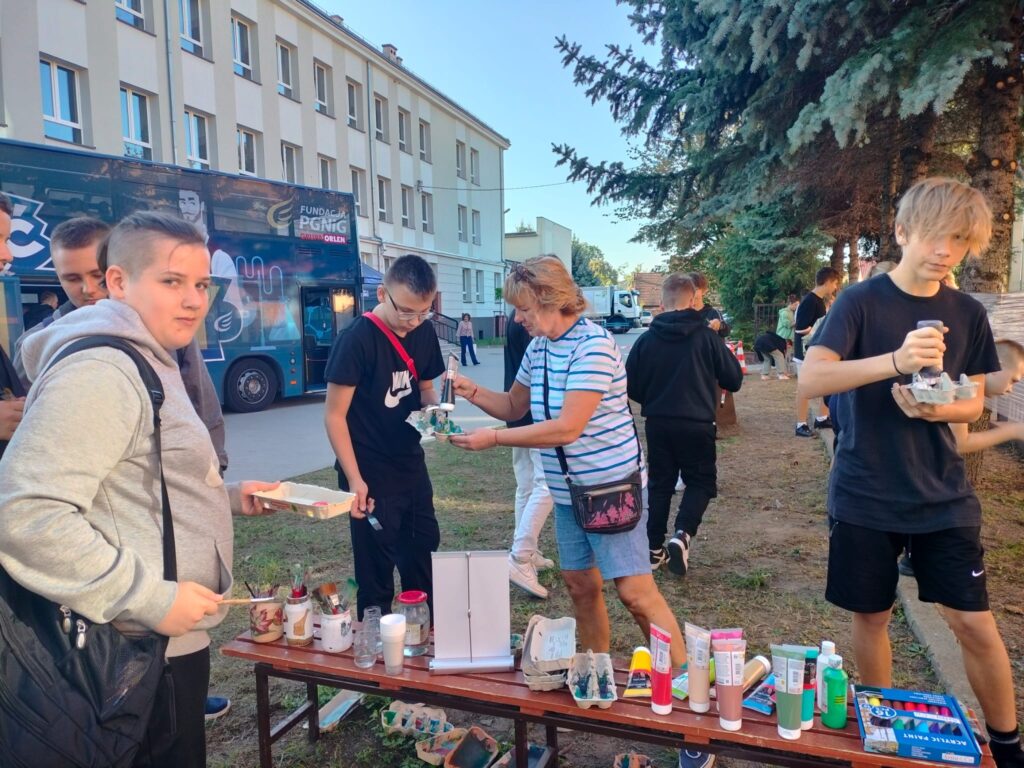 Podwórko szkolne, dzieci stoją w pobliżu folii, rozciągniętej między drzewami, po której malują pędzlami. Są w ruchu, bo biorą farby i pędzle. Pomiędzy nimi animator.