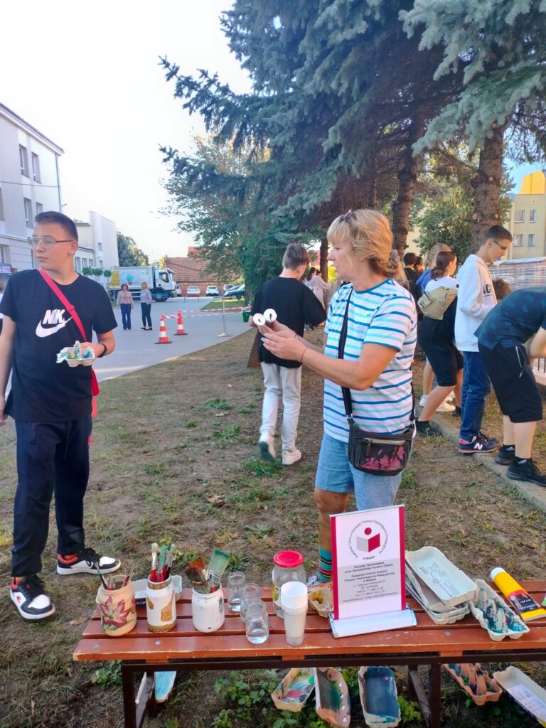 Podwórko szkolne, dzieci stoją w pobliżu folii, rozciągniętej między drzewami, po której malują pędzlami. Na pierwszym planie uczeń i nauczyciel, który daje instrukcję.