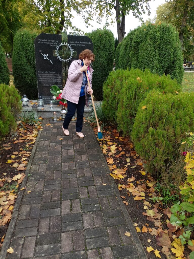 W tle wysoki pomnik na cmentarzu. Pomnik w postaci dwóch pionowych płyt z krzyżem pośrodku i cierniową koroną. Na dróżce prowadzącej do pomnika stoi kobieta z miotłą porządkująca zieleń dookoła.