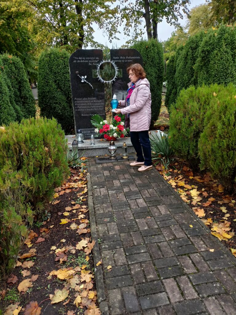 W tle wysoki pomnik na cmentarzu. Pomnik w postaci dwóch pionowych płyt z krzyżem pośrodku i cierniową koroną. Przed pomnikiem stoi kobieta zapalające szklany znicz. Po prawej i lewej stronie zielony żywopłot.

