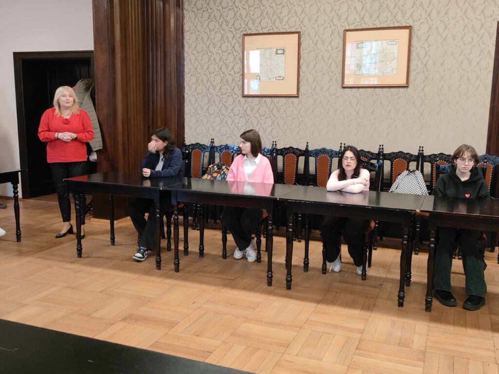 3. Na zdjęciu po lewej stronie stoi kobieta w czerwonej bluzce i czarnych spodniach. Przy stole siedzi trójka uczennic.