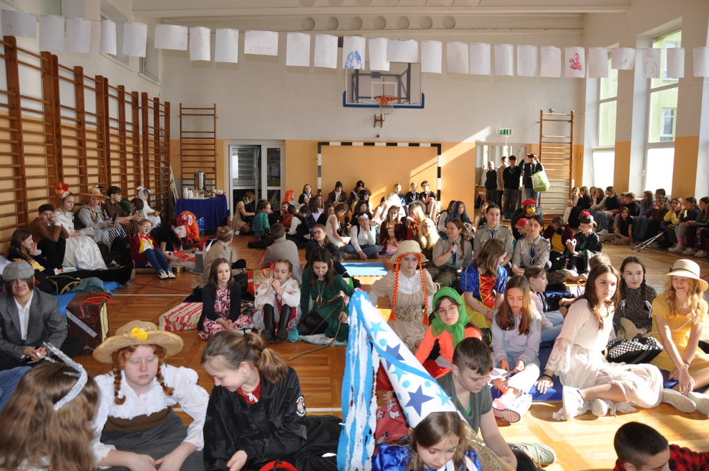 Sala, na środku siedzą dzieci w kolorowych strojach.