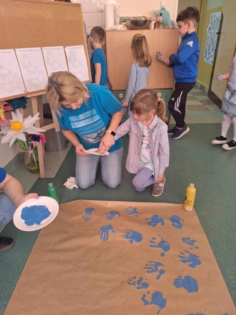 Sala lekcyjna, na podłodze szary papier, na którym są odbite niebieskie dłonie. Kobieta podaje farbkę a dziecko moczy w niej dłoń.