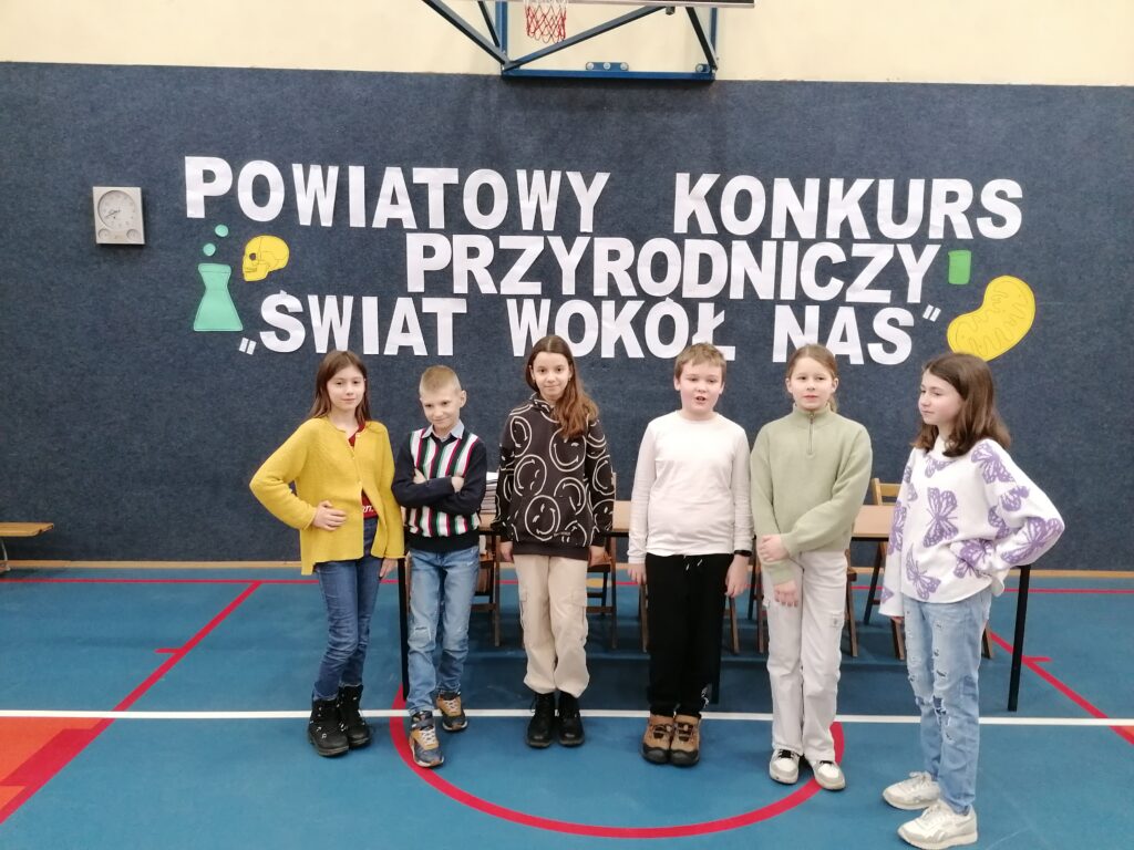 
Granatowa ściana na niej biały napis Powiatowy Konkurs Przyrodniczy „Świat wokół nas”. Na tle napisu stoi grupa uczniów