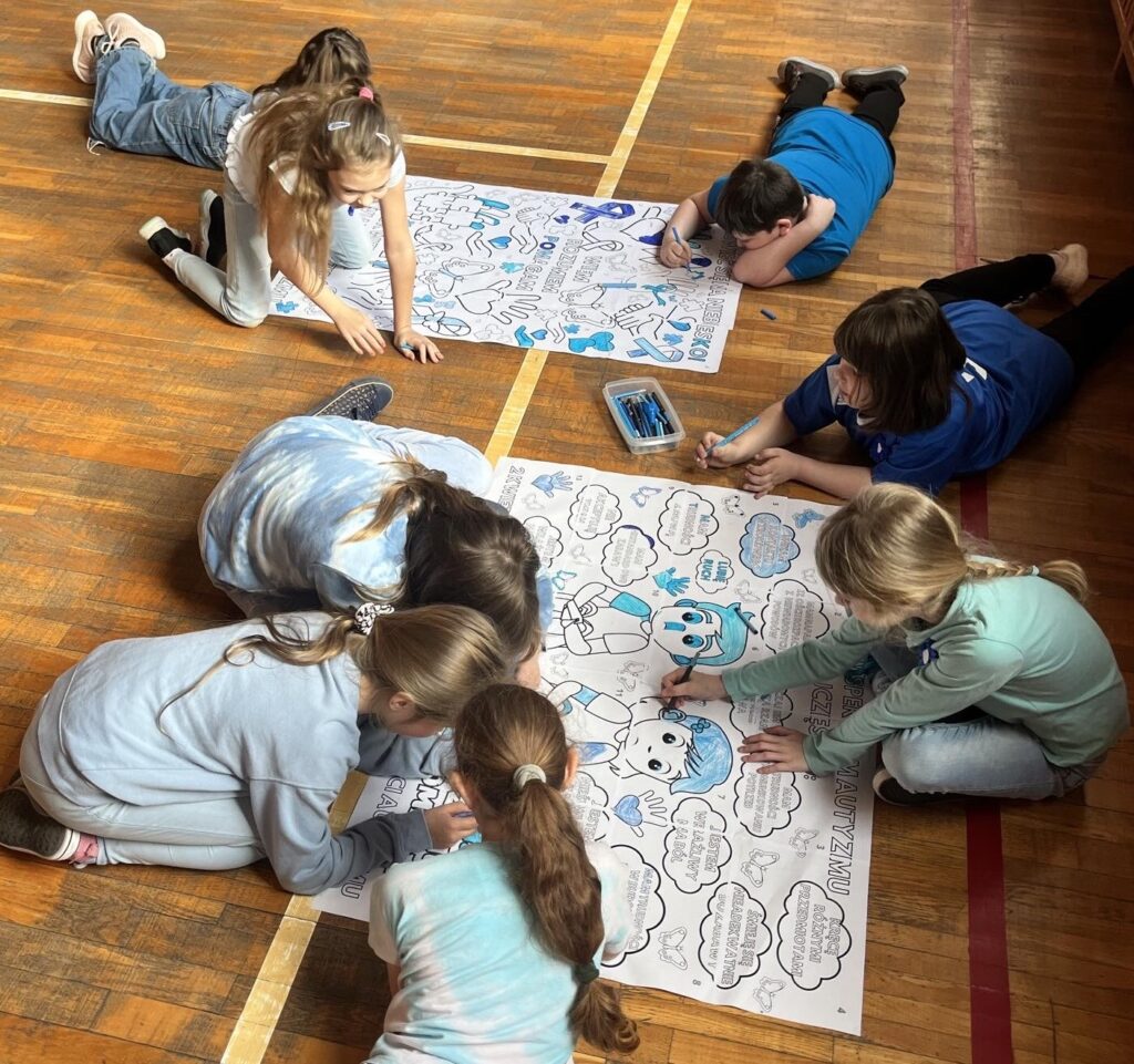 Na podłodze leżą dwa plakaty wokół leżą dzieci i wypełniają obrazki niebieskimi kolorami.