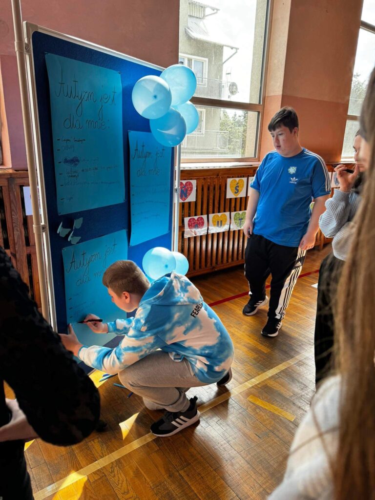 Przed niebieską tablica z niebieskimi balonami stoją dzieci. Coś piszą.