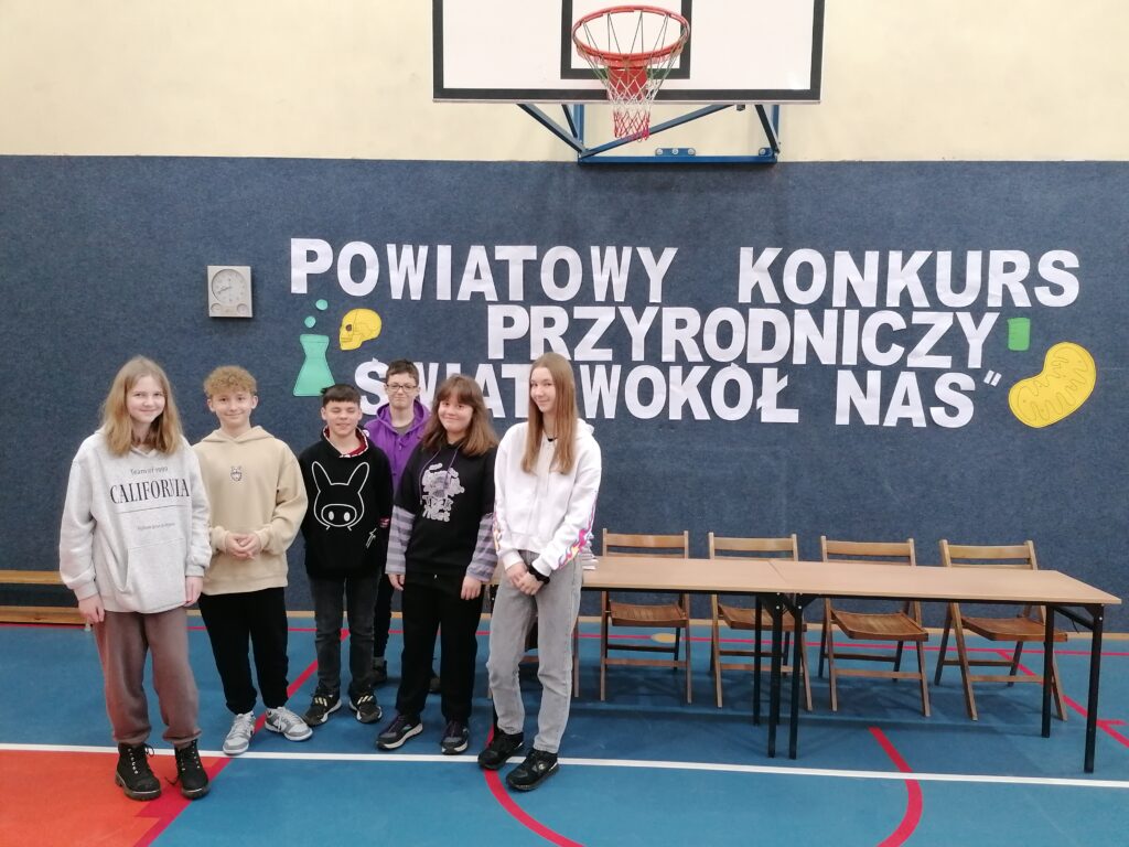 Na tle napisu Powiatowy Konkurs Przyrodniczy „Świat wokół nas” stoi grupa uczniów w różnym wieku.