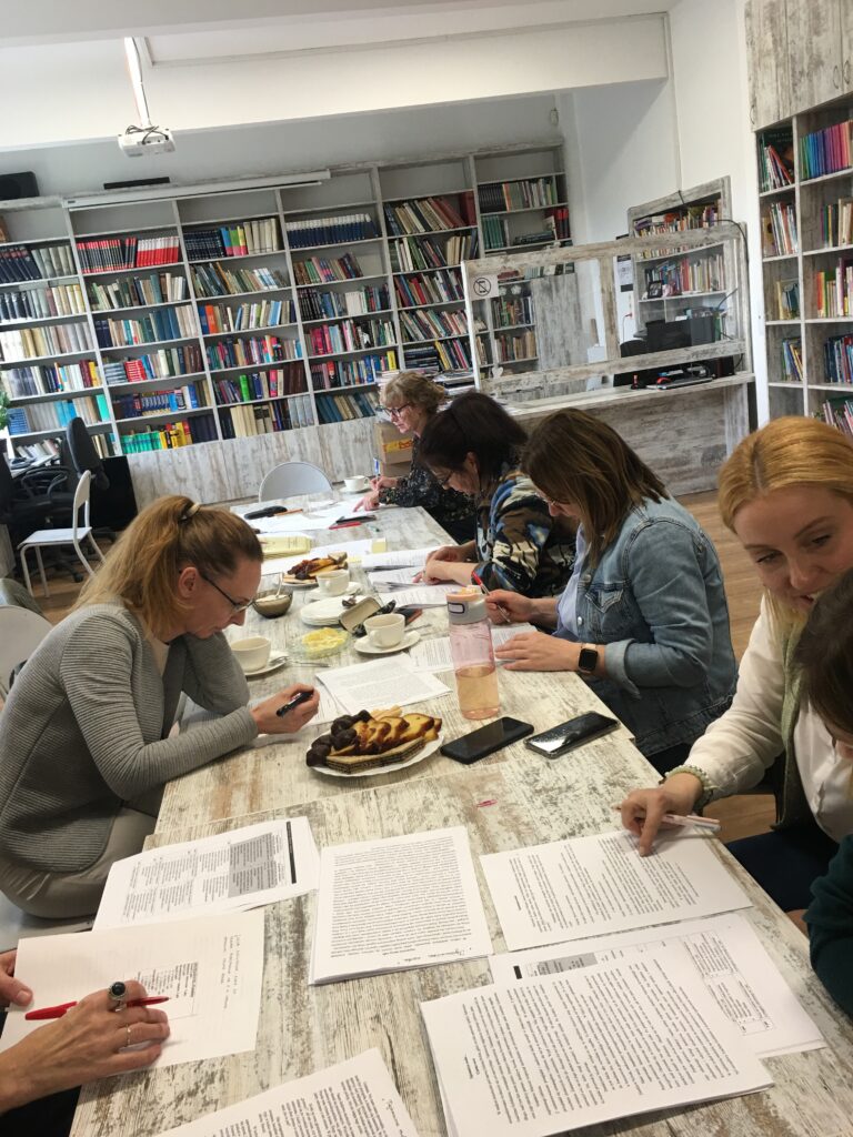 Sala biblioteczna, na środku stoły. Przy stolikach siedzą kobiety. Rozmawiają.