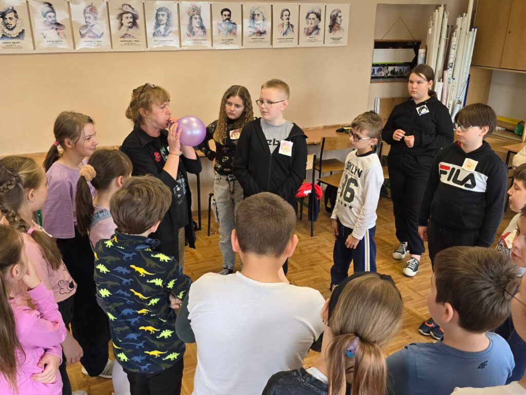 Sala lekcyjna. Na środku stoją uczniowie twarzami do siebie. W środku nauczyciel prowadzący zajęcia dmucha w balon..