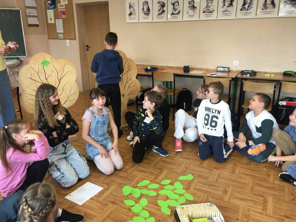 Sala lekcyjna. Uczniowie siedzą na podłodze, tworzą krąg. Na podłodze leżą zielone listki wykonane z papieru. Za nimi dwa duże drewniane drzewa. Dzieci do nich podchodzą i coś naklejają. 