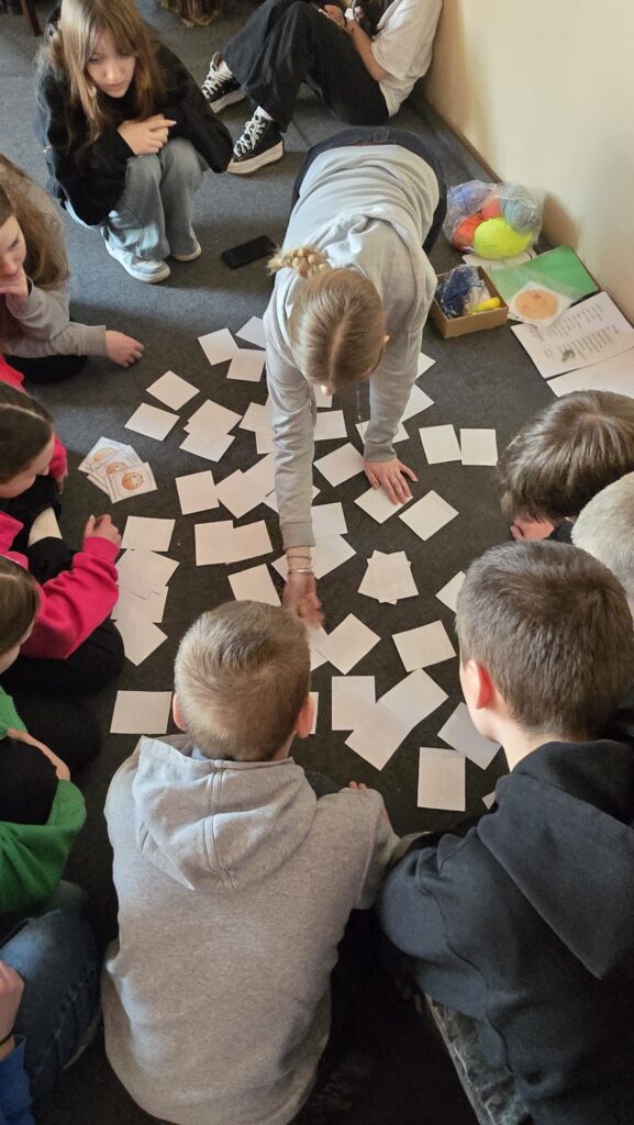 Aula szkolna, na środku sceny uczniowie pochylają się nad rozrzuconymi kartkami. 