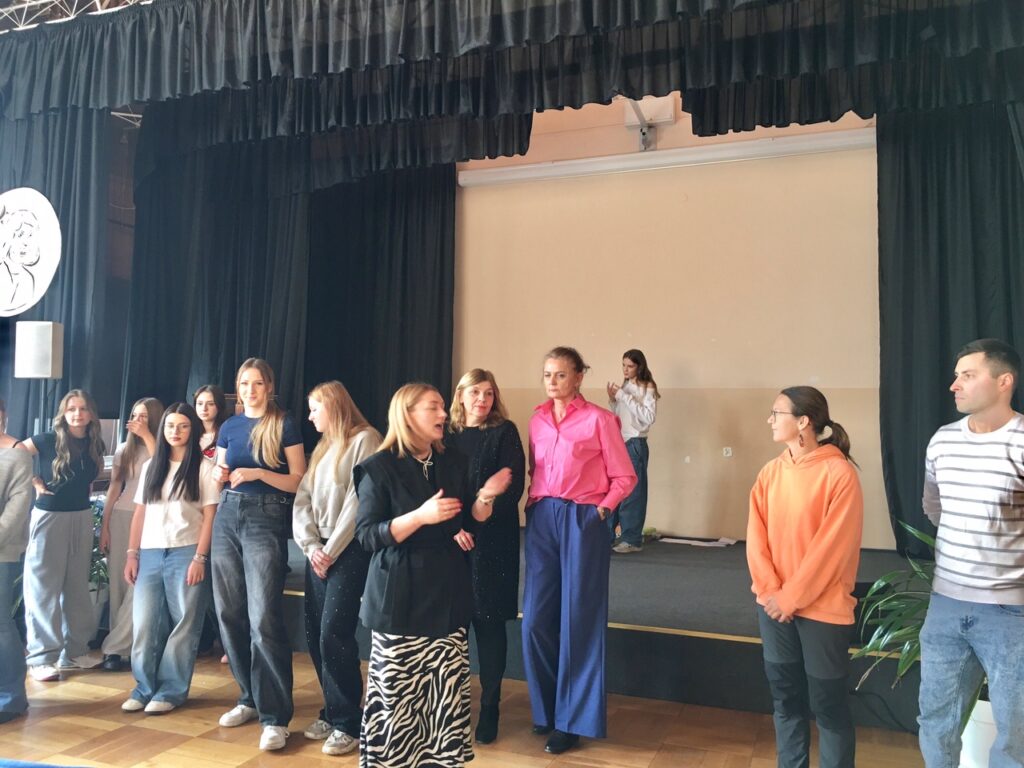 Aula szkolna, na środku kobiety, które mówią coś do uczestników.
