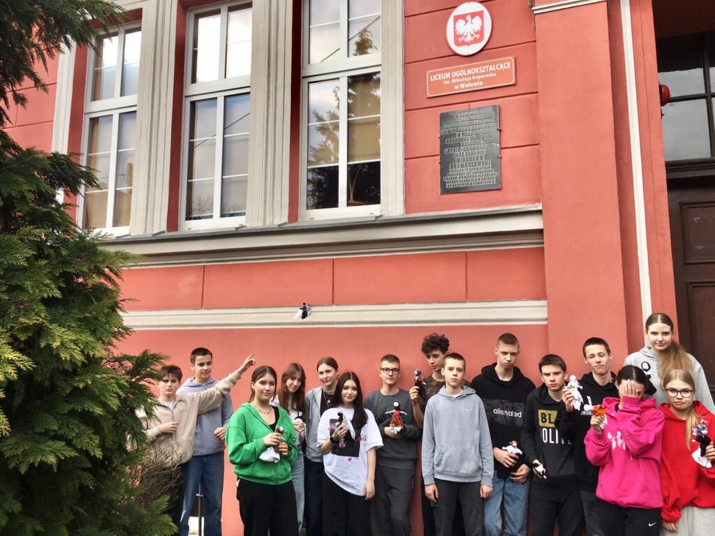 Grupa uczniów przed budynkiem szkoły. Młodzież stoi na tle różowej ściany, mają w ręku szmaciane laleczki.