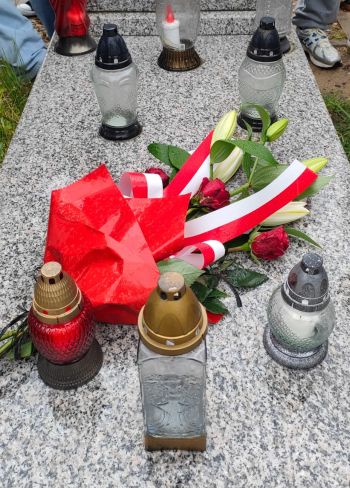 Wiązanka kwiatów z biało-czerwona wstążką leży na płycie grobu obok zniczy,