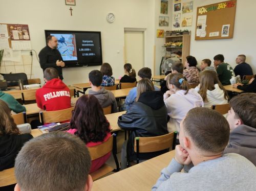 Uczniowie siedzą w ławkach, nauczyciel stoi przy tablicy interaktywnej