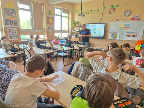 Uczniowie siedzą w ławkach, nauczyciel stoi przy tablicy interaktywnej.