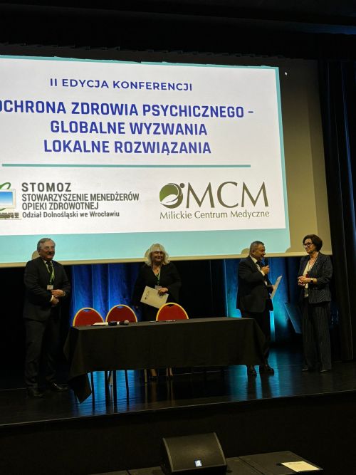 na środku sceny wyświetlana jest prezentacja . Stoją 4 osoby dwie kobiety i dwóch mężczyzna. Jeden z nich trzyma mikrofon i mówi do publiczności. 