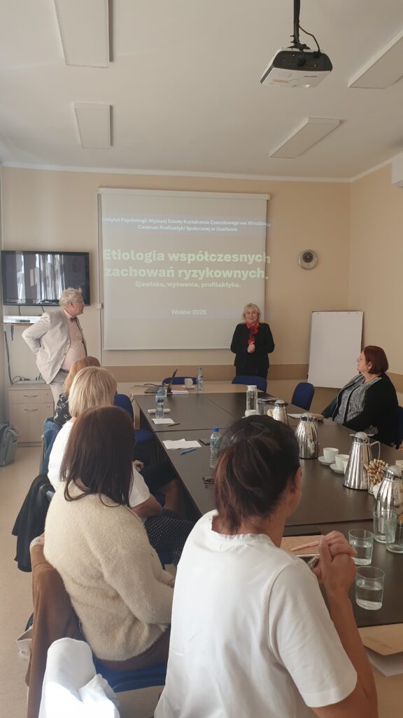 w sali szkoleniowej wisi ekran na którym wyświetlana jest prezentacja. Na środku stoi kobieta i przemawia. Po lewej stronie stoi mężczyzna. Po środku stół przy którym siedzą uczestnicy szkolenia