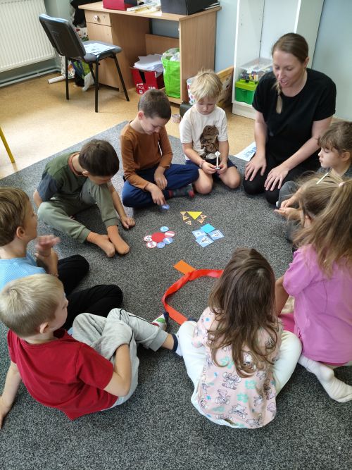 Dzieci z panią siedzą w kole na dywanie i wykonują zadanie.