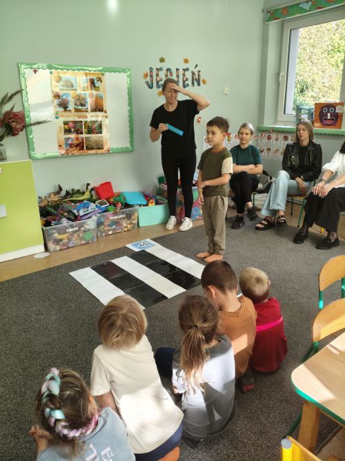 Dzieci z panią siedzą na dywanie. Przy białej ścianie stoi nauczycielka i wskazuje ręką.
Przed dziećmi stoi chłopiec przechodzący przez pasy.
