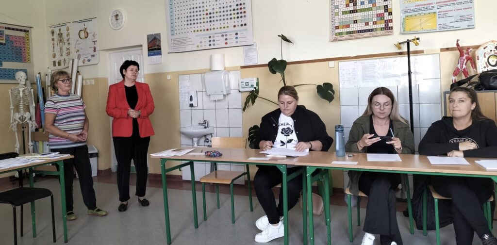 Klasa, w której przy stolikach siedzą nauczyciele. Na stolikach leżą kartki, notatniki. Dwie kobiety stoją przed siedzącymi przy stolikach osobami.