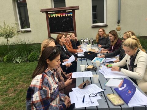 Zielona klasa na świeżym powietrzu, na środku stoły. Wokół siedzą nauczyciele. Na stole leżą kartki, notesy oraz stoją talerzyki z ciastem i kubki i termosy.