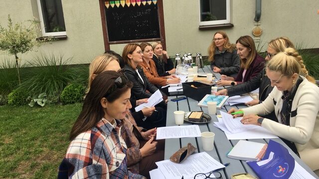 Zielona klasa na świeżym powietrzu, na środku stoły. Wokół siedzą nauczyciele. Na stole leżą kartki, notesy oraz stoją talerzyki z ciastem i kubki i termosy.