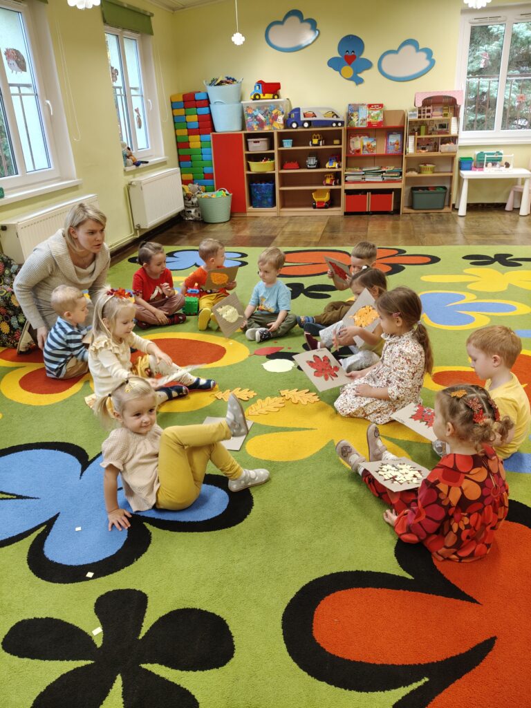 Dzieci z siedzą w kole na kolorowym dywanie. Przed sobą maja prace plastyczne – wyklejone liście.
Pani jest wśród dzieci z głową skierowaną w ich stronę.
W tle dekoracja na ścianie: dwie chmurki i ptaszek.
