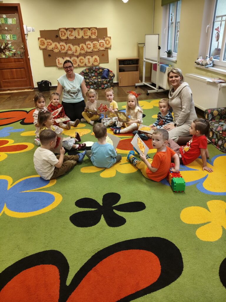 Dzieci z siedzą w kole na kolorowym dywanie głowy skierowane w stronę fotografującego.
Dwie panie siedzą wśród dzieci.
W tle dekoracja na ścianie napis Dzień spadającego liścia.